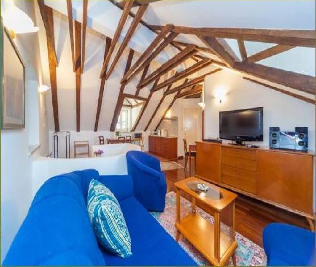 A modern living room with a high ceiling and exposed wooden beams, a blue sofa, a wooden coffee table, and a flat-screen TV.