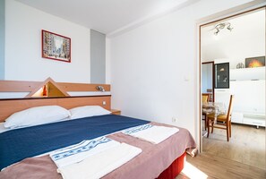 Ein Hotelzimmer mit Bett, Holz-Kopfstütze, einem Gemälde an der Wand und einem Essbereich, der durch eine offene Tür sichtbar ist.
