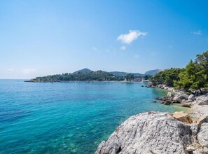A coastal landscape with clear turquoise waters, rocky shores, and a small island with buildings in the distance.