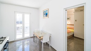 Een kleine, lichte kamer met een tafel met glazen blad, een witte stoel en een bed met een gele en oranje dekbed.