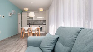 A modern living room with a grey sofa, a dining area with white chairs, and a kitchen with white cabinets and a hanging light fixture. - Dva Galeba - Standard One Bedroom Apartment with Terrace (101) (Bol)