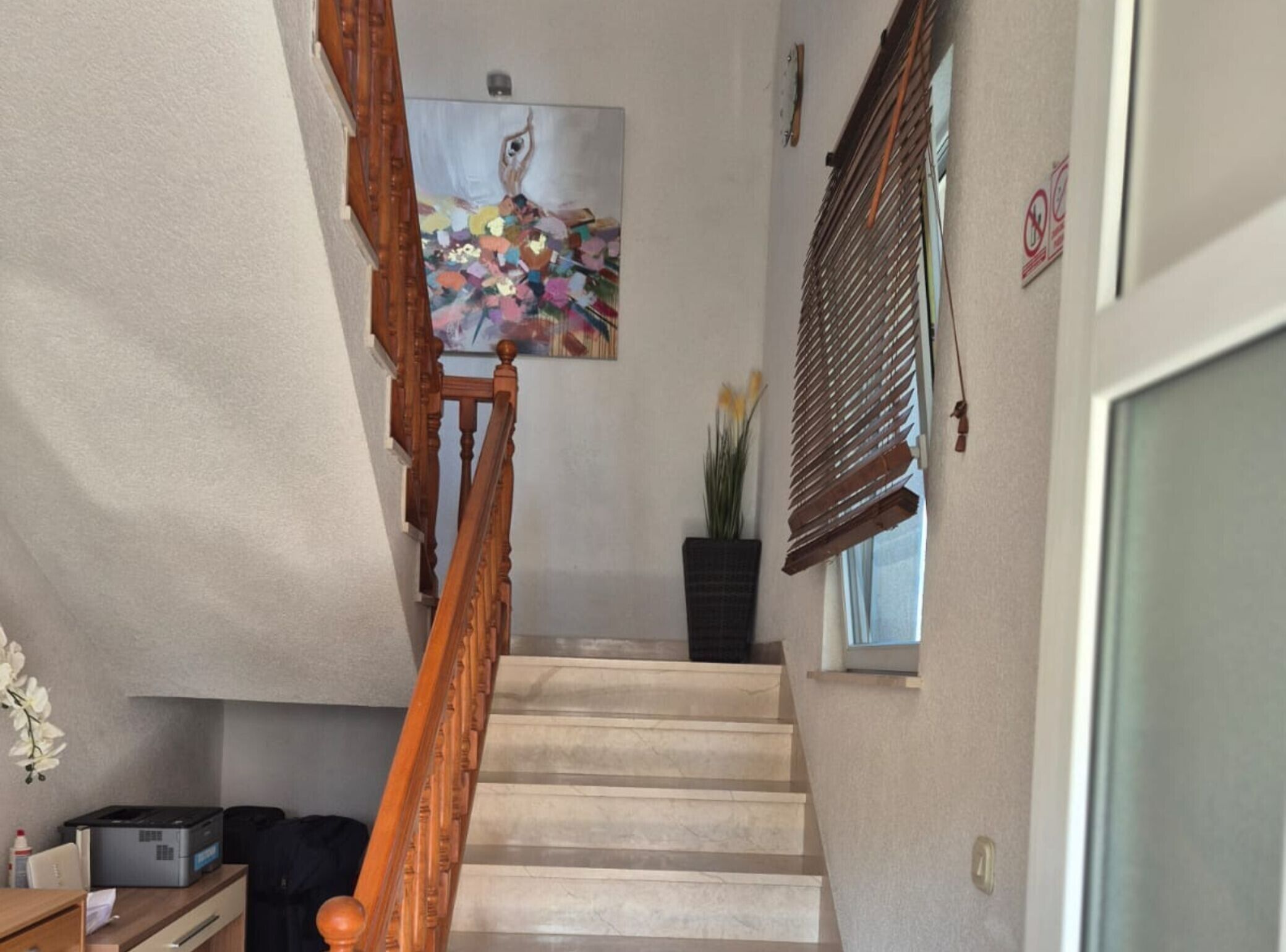 Una escalera con barandilla de madera, un cuadro en la pared, una planta en maceta y una ventana con persianas.