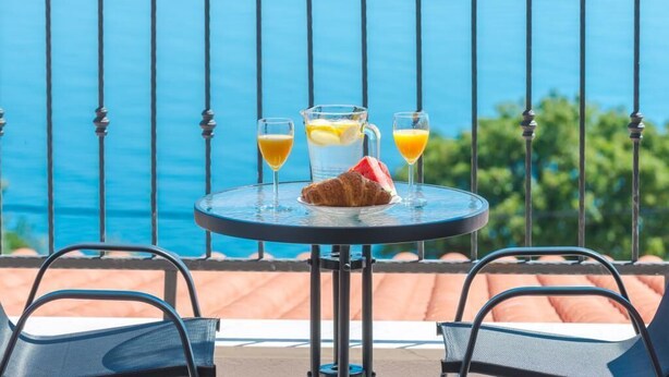 A table with a croissant, a pitcher of lemonade, and two glasses of orange juice on a balcony with a view of the sea.