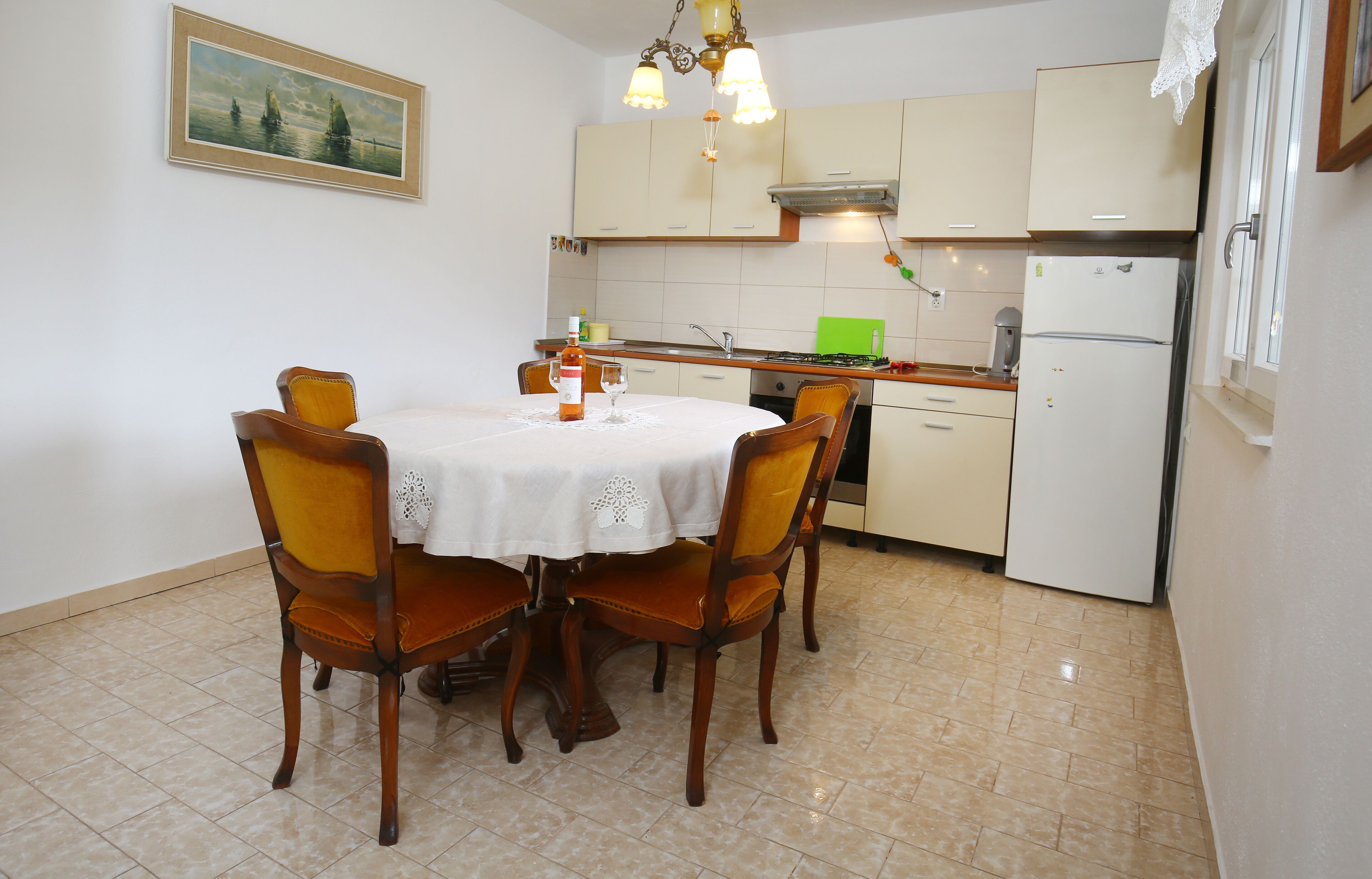A dining area with a round table, four chairs, a chandelier, and a kitchen in the background.