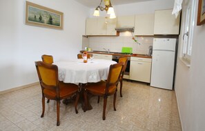 Uma área de refeições com uma mesa posta para quatro pessoas, uma cozinha com armários e eletrodomésticos, e um quadro na parede.
