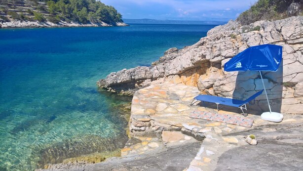 A stone platform with a blue umbrella and a beach chair overlooking a clear blue sea.