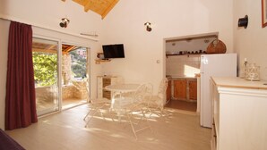 A spacious living area with a sliding glass door leading to a balcony, a round dining table with chairs, a television mounted on the wall, and a kitchen area with cabinets and a refrigerator.