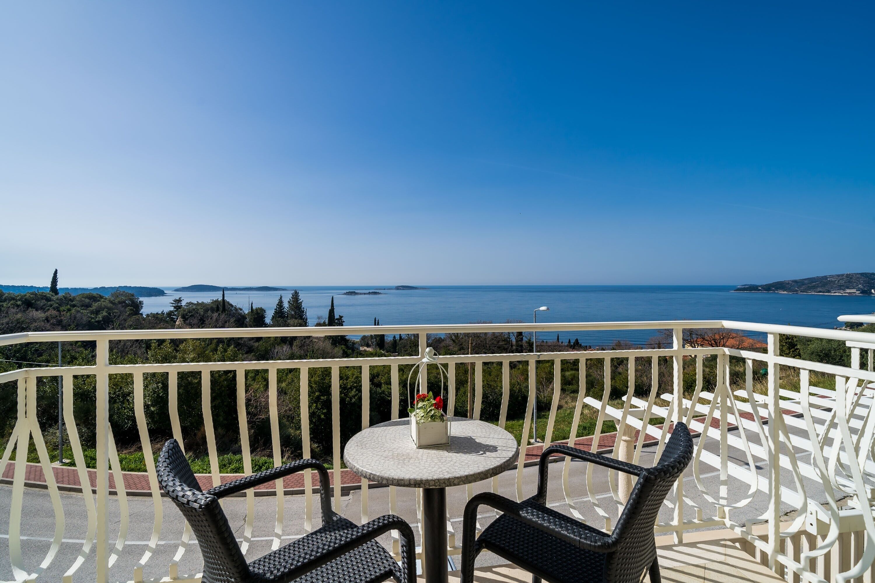 Uma varanda com cadeiras de vime e uma mesa redonda, com vista para o mar e o litoral ao longe.