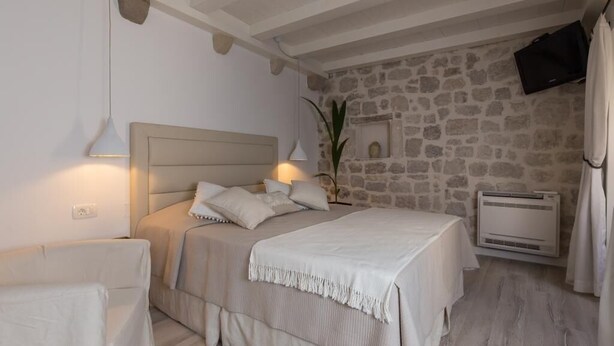 A bedroom with a stone wall, a bed with white bedding, a bedside table, a lamp, and a mounted TV.