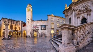 A historic square with a prominent clock tower, a fountain, and classical architecture.