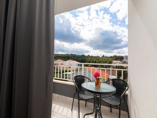 A balcony with a round glass table and two chairs, a potted plant, and a view of a hilly landscape with buildings.