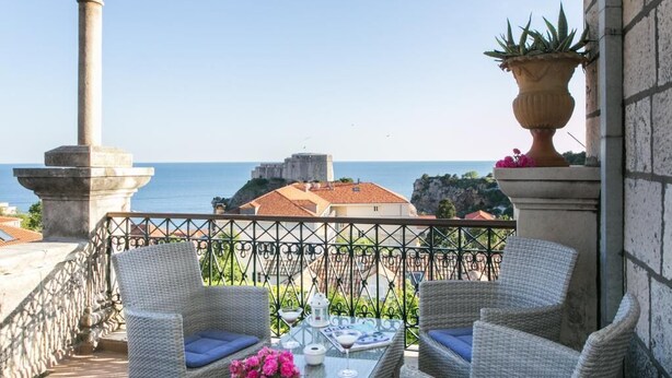 Un balcón con sillas de mimbre, una mesa de vidrio y vistas al mar y edificios.