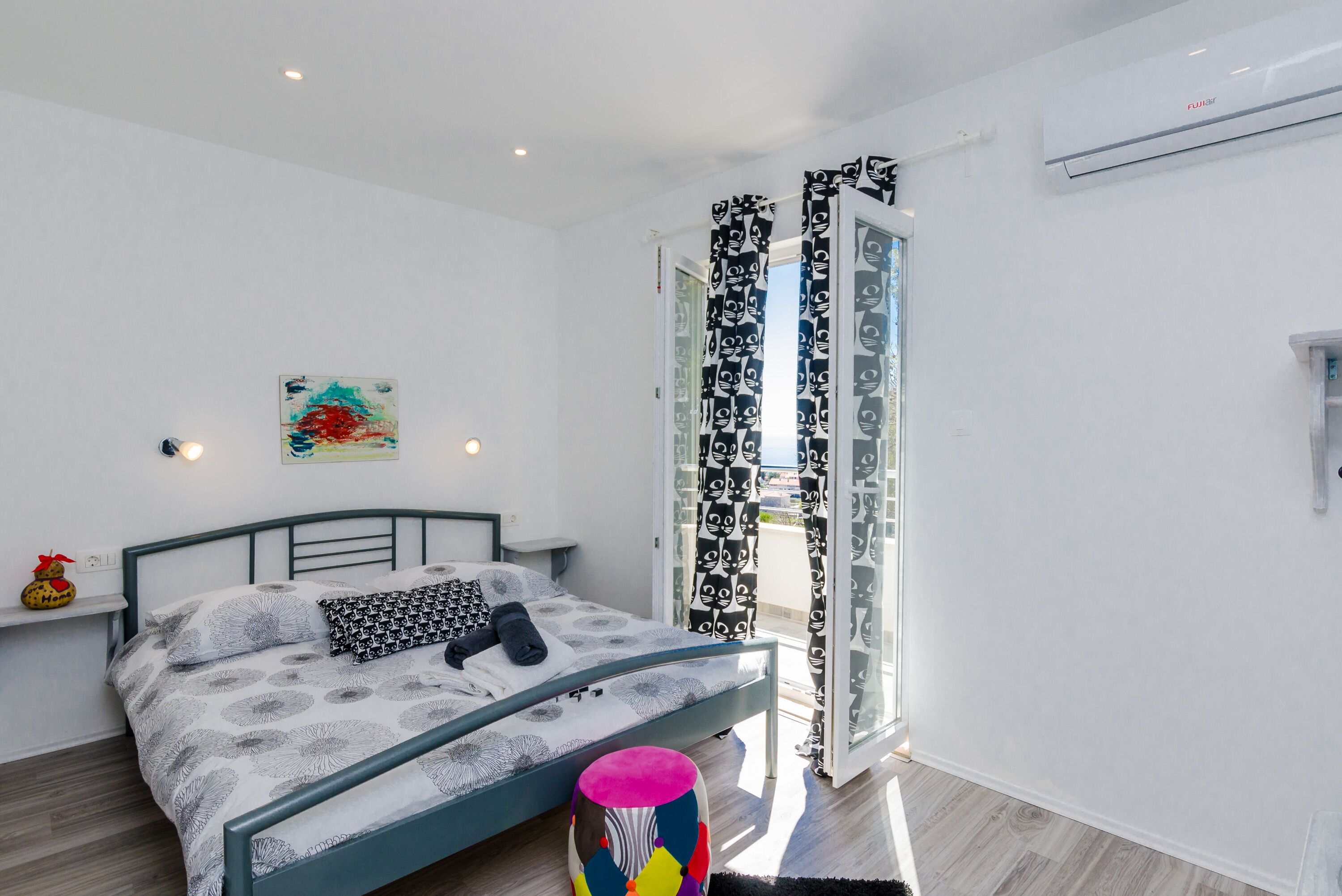 Un dormitorio con cama, una mesita de noche, un taburete de colores, una ventana con cortinas estampadas y un aire acondicionado instalado en la pared.