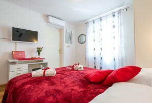 Une chambre avec une couvre-lit rouge, du linge de lit blanc, un téléviseur fixé au mur, un bureau avec une boîte rouge et un vase de fleurs, et une fenêtre ornée de rideaux à motifs floraux.
