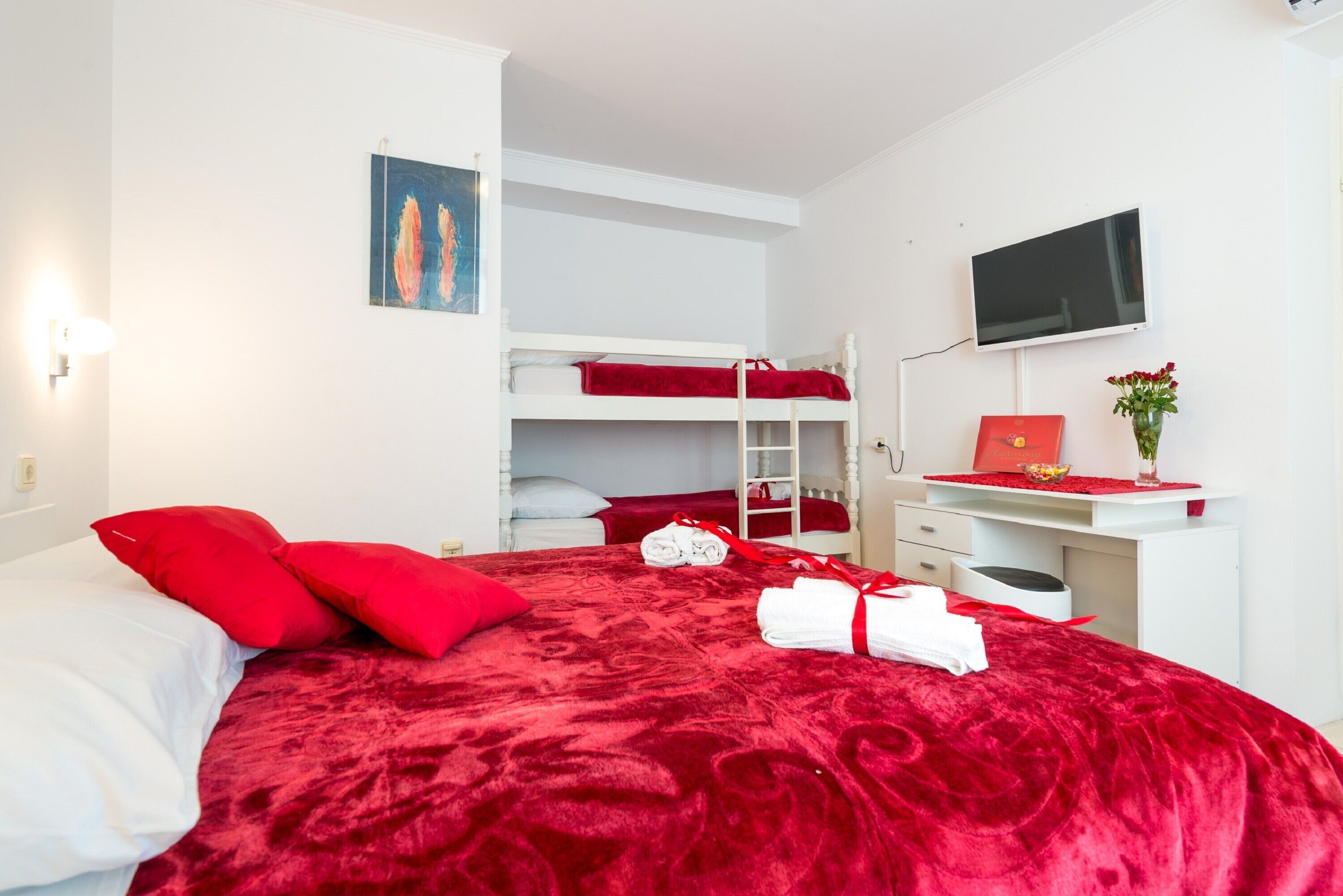 Una habitación con una cama grande, una litera, un escritorio con una computadora y un televisor en la pared.