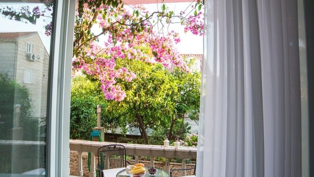 A balcony with a table and chairs, a window with white curtains, and a view of a tree with pink flowers.