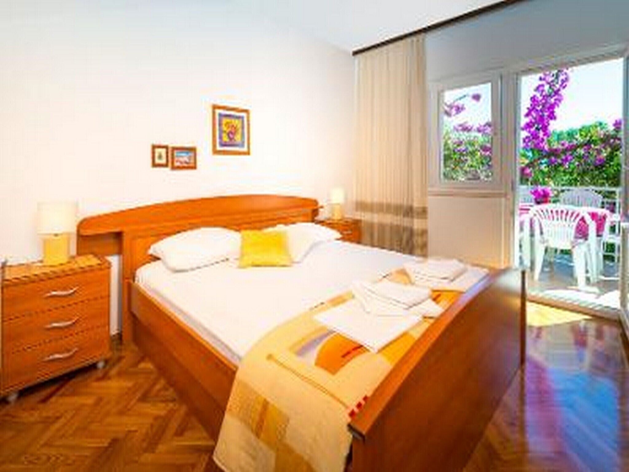 Een hotelkamer met een houten bed, een nachtkastje met een lamp, een raam met uitzicht op paarse bloemen, en een balkon met een witte stoel.