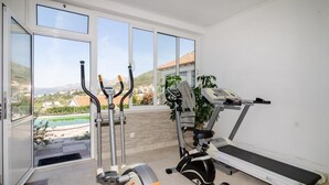 Un gimnasio en casa con cinta de correr, elíptica y bicicleta estática, ventanas amplias con vista a una piscina y colinas.