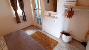 Un dormitorio con cama, ventana con cortinas, espejo, estante con una planta y puerta que da a otra habitación.