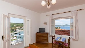 Una habitación con un aparador de madera, un televisor, una mesa de comedor con sillas, una alfombra roja y vistas al mar y las colinas a través de una ventana.