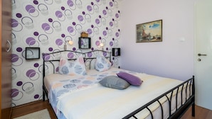 A bedroom with a patterned wallpaper, a bed with multiple pillows, a nightstand, a framed picture on the wall, and a door to the right.