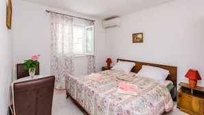 A bedroom with a bed, a nightstand, a table with a chair, a window with curtains, and an air conditioning unit. - Villa Bind Comfort Double Room,Patio&Garden ViewR4 (Obod)