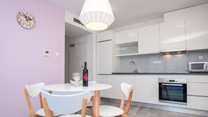 A modern kitchen with white cabinets, a built-in oven, and a microwave. There is a dining table with chairs and a wine bottle on it.