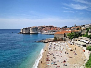 A coastal town with a historic fortification, a sandy beach with umbrellas, and clear blue waters.