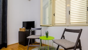 A room with a wooden floor, a television on a dark cabinet, two patterned chairs, a small green table with a plant, and a window with blinds. - The Point Room - Double Room with City View (Dubrovnik)