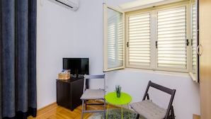 A small room with a TV, a desk, two chairs, a small table with a plant, and a window with blinds. - The Point Room - Double Room with City View (Dubrovnik)