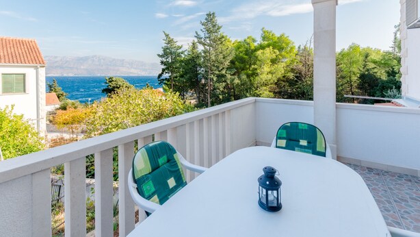 A balcony with a white table and chairs, overlooking a coastal view with trees and a building in the background. - Villa Stella - Two Bedroom Apartment with Terrace and Sea View (Stella) (Splitska)