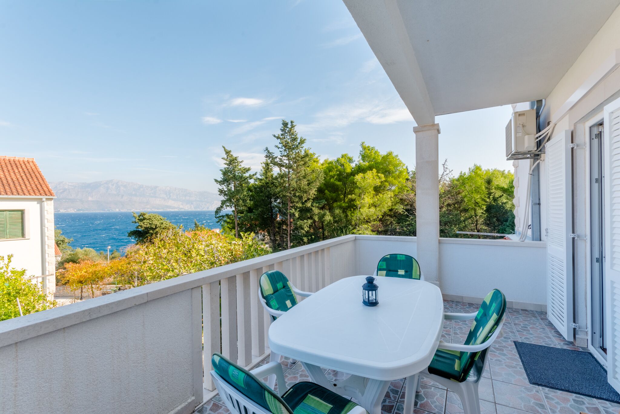Een balkon met een witte tafel en groene stoelen, met uitzicht op de kust, bomen en gebouwen.