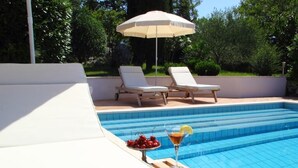 Ein Swimmingpool mit zwei weißen Liegestühlen und einem Sonnenschirm, eine Ablage mit Kirschen und einem Cocktail, sowie ein gefliestes Deck.