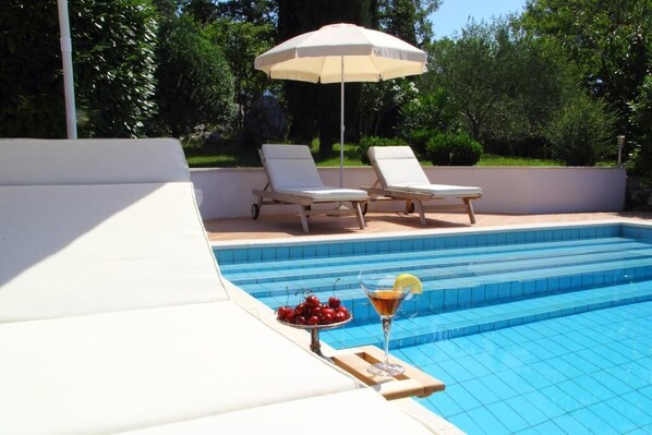 A swimming pool with two white lounge chairs and an umbrella, a tray with cherries and a cocktail, and a tiled deck.