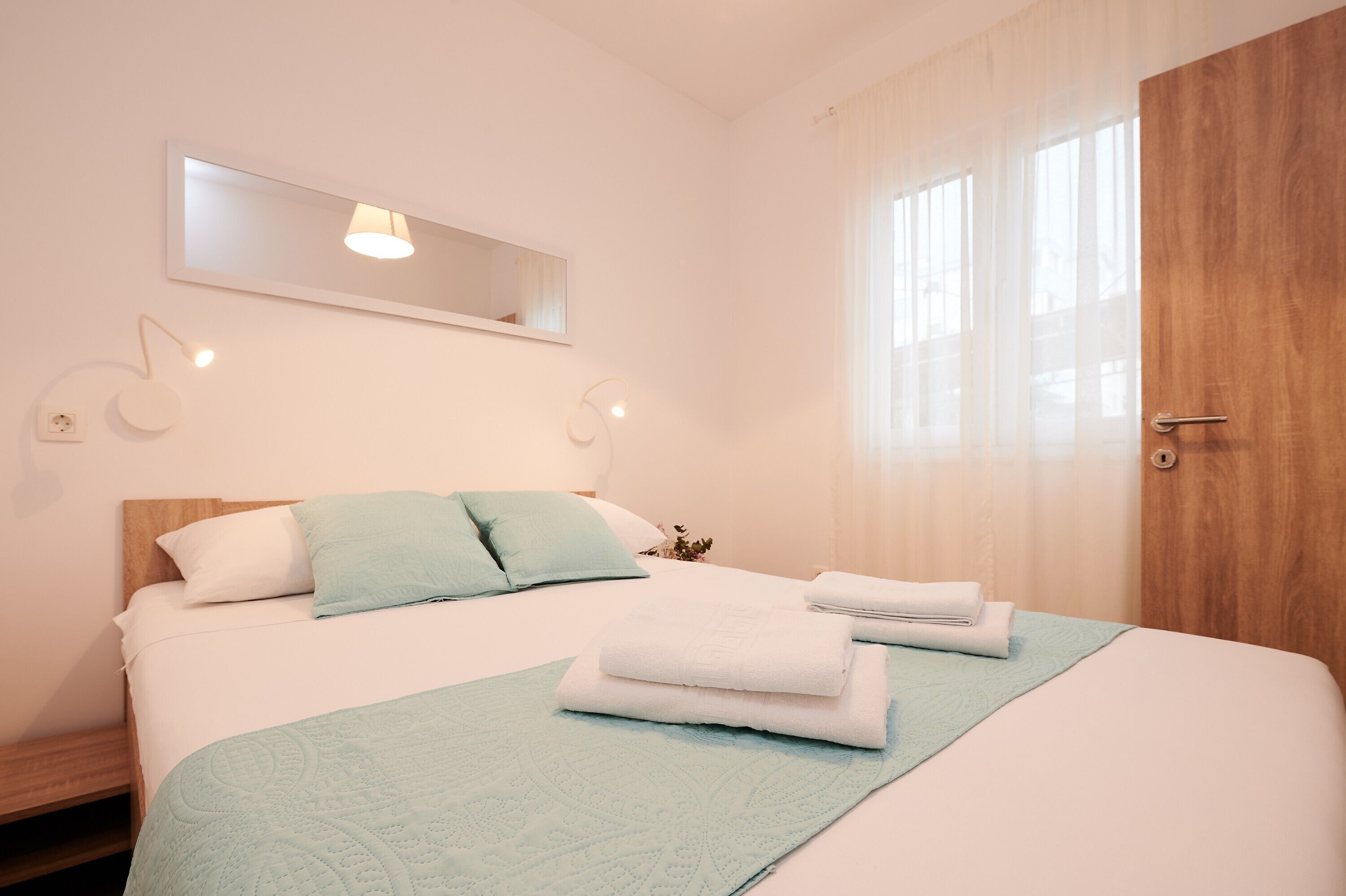 Ein ordentlich bezogenes Bett mit weißen Laken und einer hellblauen Tagesdecke, eine Holztür und ein großes Fenster mit transparenten Vorhängen.