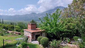A rustic stone building with a tiled roof, surrounded by lush greenery and a clear view of distant mountains.