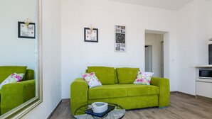 Un sofá verde con cojines estampados, una mesa de centro de cristal con un libro y un control remoto, y un reloj de pared.