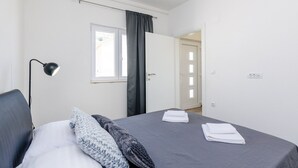 Un dormitorio con cama, lámpara de noche, ventana con cortina y puerta que conduce a otra habitación.