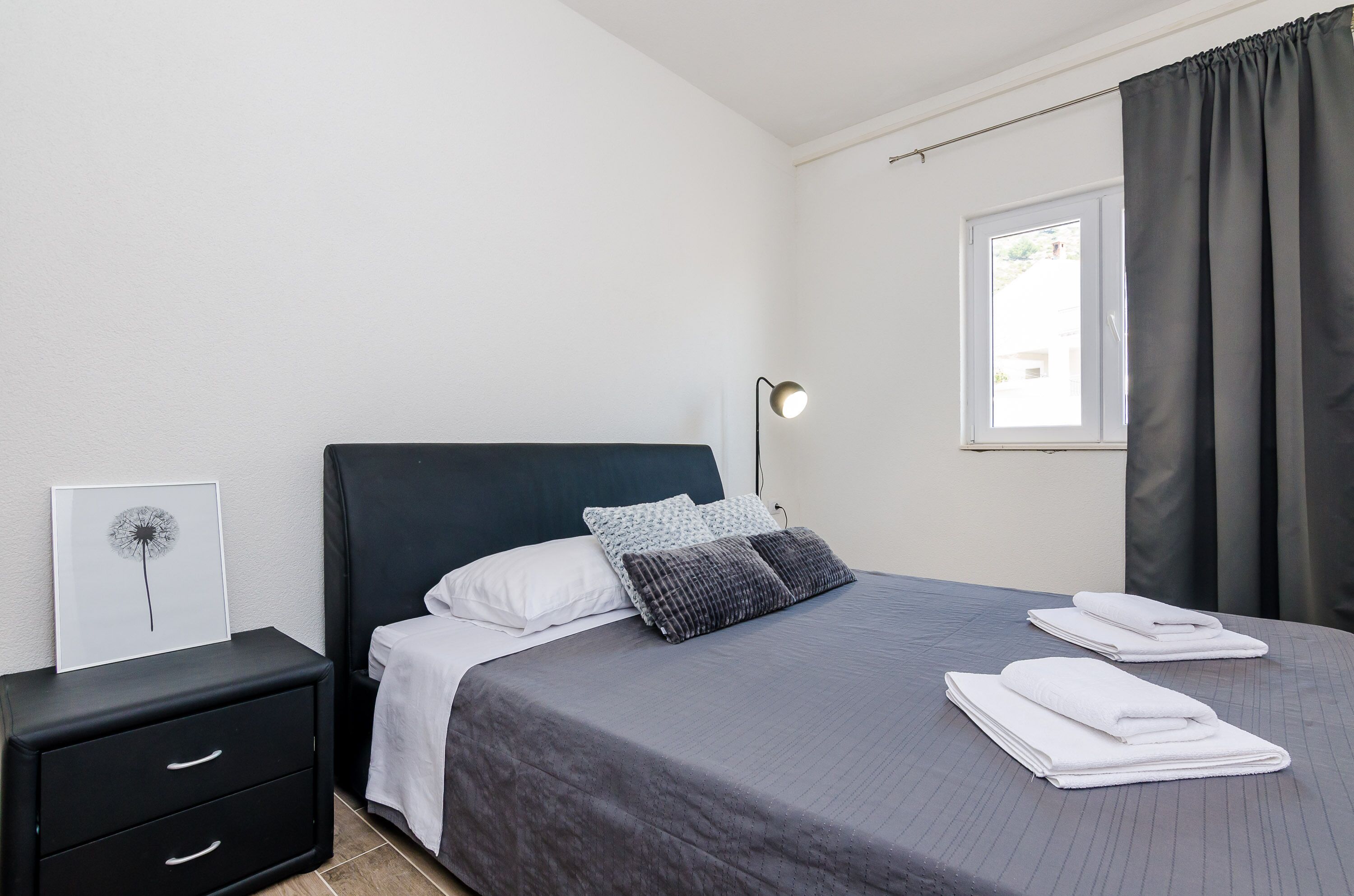 Een netjes opgemaakt bed met een grijze deken en twee kussens, een nachtkastje met een lamp, en een ingelijst beeld aan de muur.
