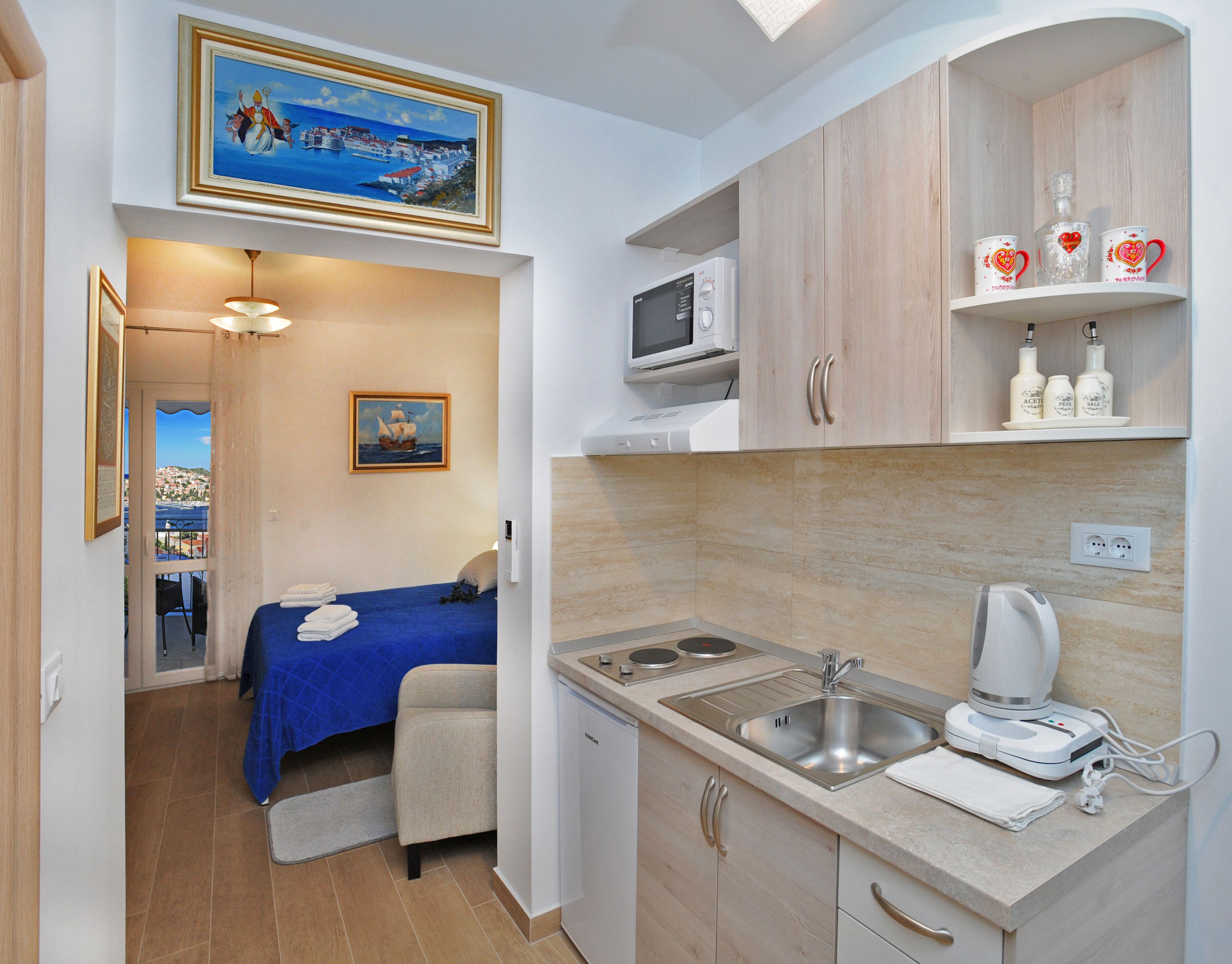 Habitación de hotel compacta con cama, una kitchenette pequeña, microondas, lavabo y cafetera. Hay un cuadro en la pared y vista al mar por la ventana.