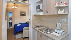Une chambre d’hôtel compacte comprenant un lit, une petite kitchenette, un four à micro-ondes, un évier et une machine à café. Un tableau orne le mur et la fenêtre offre une vue sur la mer.