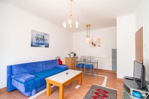 Uma sala de estar com sofá azul, mesa de centro de madeira, televisão em móvel, área de jantar com mesa e cadeiras, e cozinha com micro-ondas e chaleira.