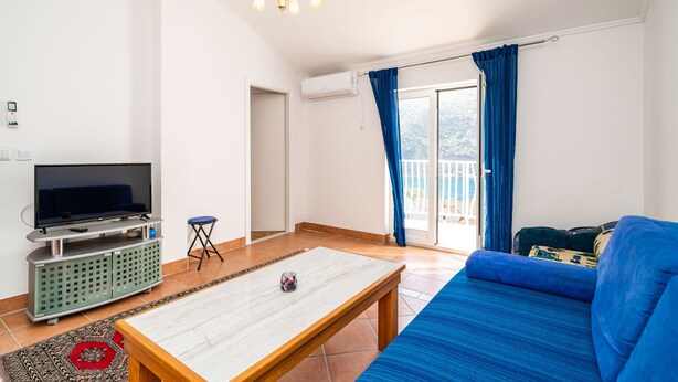 A living room with a flat-screen TV, a wooden coffee table, a blue sofa, and a balcony with blue curtains. - Apartments Peco - Studio Apartment with Balcony and Sea View (Mokosica)
