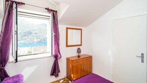 A bedroom with a bed, a wooden wardrobe, a window with purple curtains, a mirror, and a view of a body of water and buildings. - Apartments Peco - Studio Apartment with Balcony and Sea View (Mokosica)