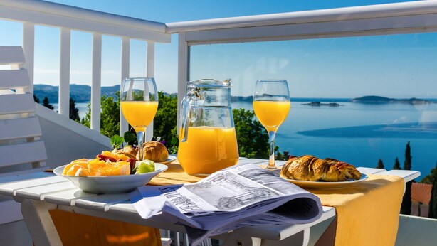 Aamiaispöytä, jolla on tarjolla croissantteja, appelsiinimehua ja sanomalehti, ja josta avautuu upea näkymä merelle ja vuorille.