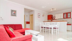 A modern living room with a red sofa, white coffee table, and a dining area with white chairs. There is a kitchen in the background with red cabinets.