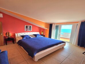 Habitación de hotel con cama, mesita de noche, un cuadro en la pared y vista al océano a través de una puerta corredera.