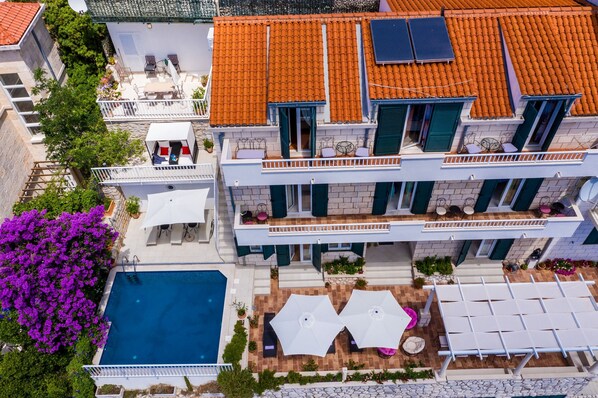 Ein mehrstöckiges Gebäude mit Ziegeldach, Außenbereich mit Sitzgelegenheiten, Schwimmbad und Garten mit violetten Blumen.