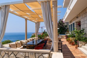 Eine Terrassensitzgruppe im Freien mit Tisch und Stühlen, eine Pergola mit Vorhängen und Blick auf das Meer.
