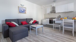 Una sala de estar moderna con un sofá gris, una mesita blanca y una cocina con armarios blancos y una mesa de comedor.